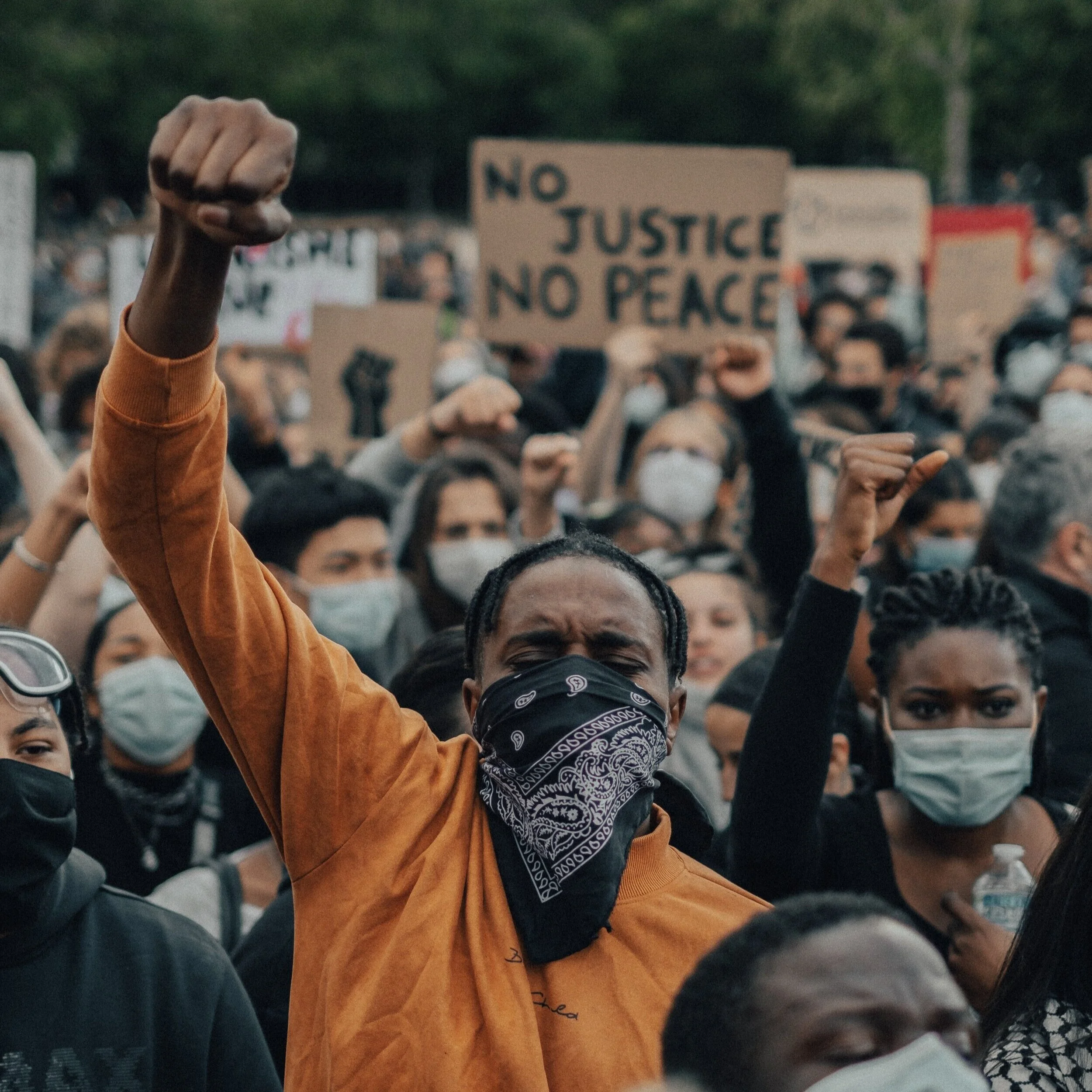 Protesters wearing masks raise fists in the air, sign in the background reads "No Justice No Peace"