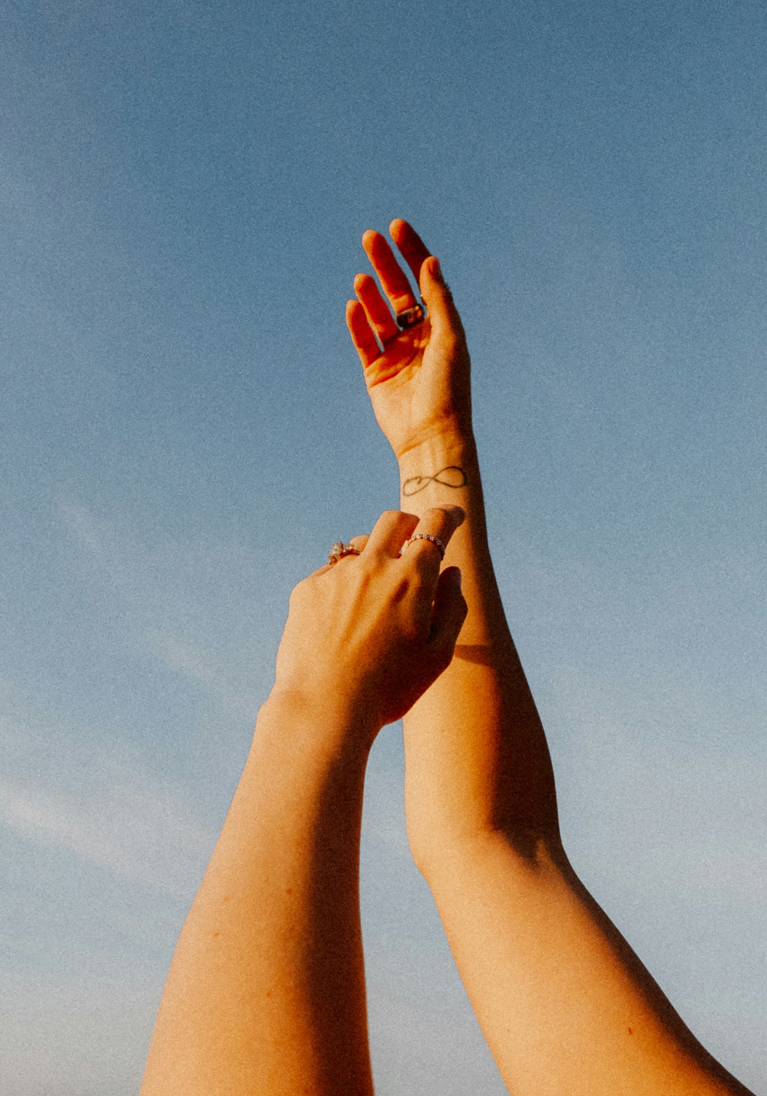 Two hands with rings and tattoos reaching towards the sky, bathed in warm sunlight, against a clear blue sky.