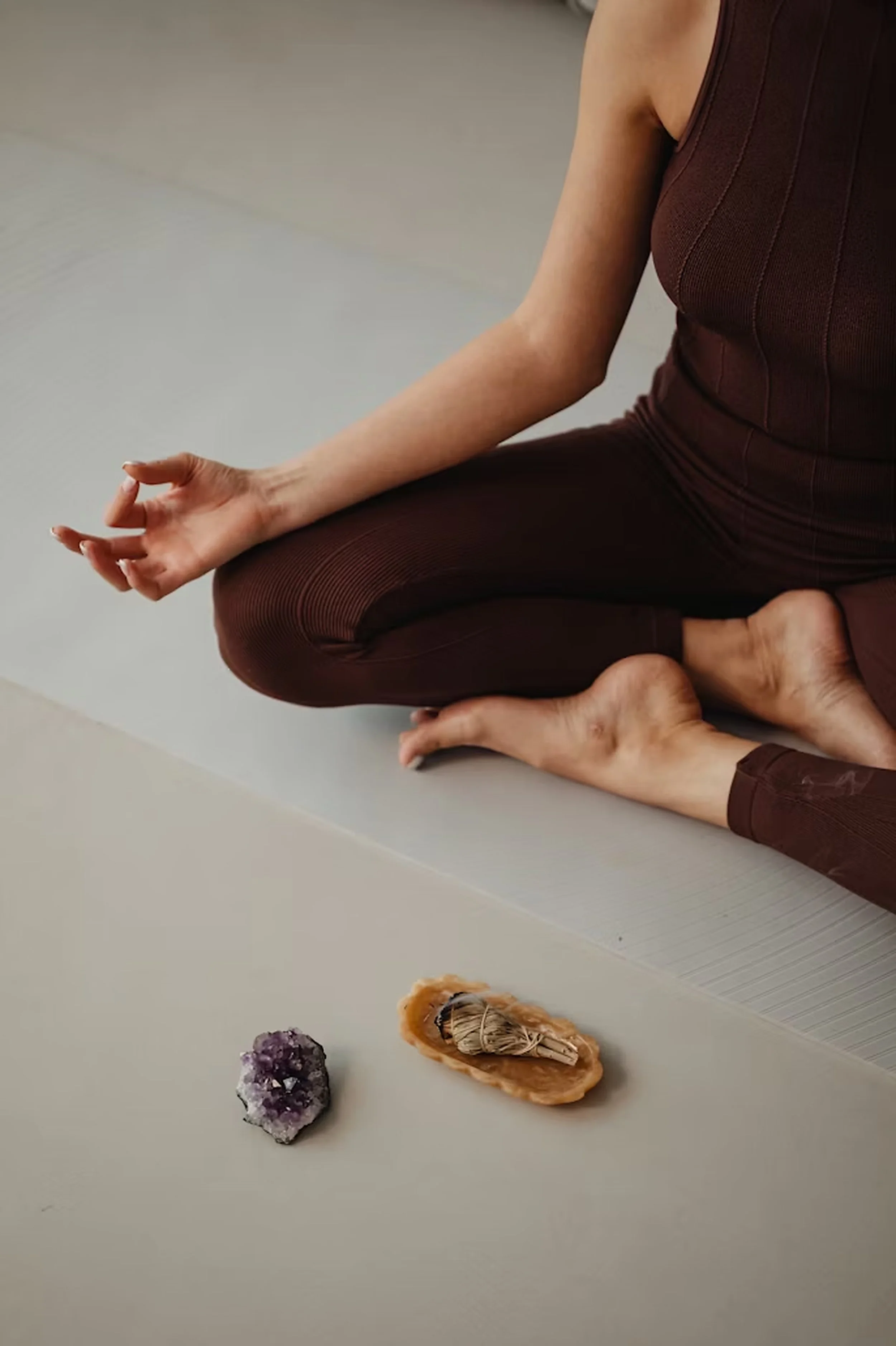 Person sitting cross-legged on the floor with a hand in a meditation gesture, wearing a brown sleeveless top and pants, with a few crystals and stones placed on a table nearby.