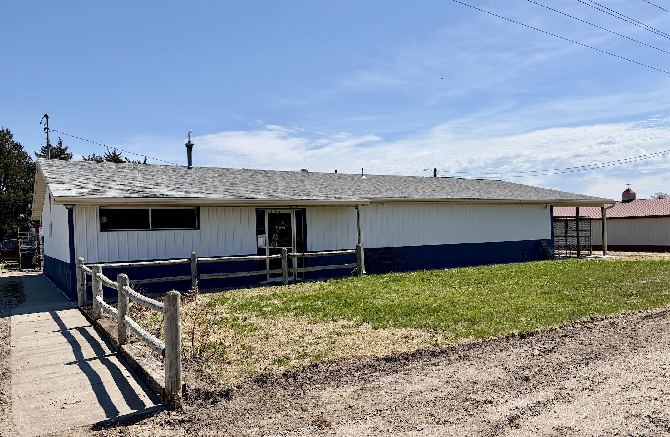 A single-story white building with a gray shingle roof, a small porch, and a grassy yard. There is a wooden fence along the sidewalk, and some trees in the background. The sky is partly cloudy with some overhead power lines visible.