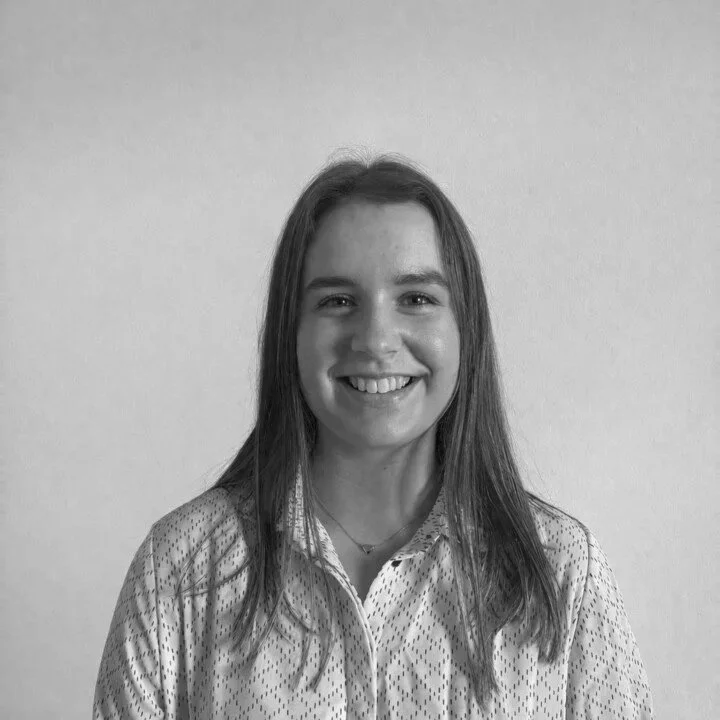 A young woman with long hair and a bright smile, wearing a collared shirt with a subtle dotted pattern, standing against a plain wall.
