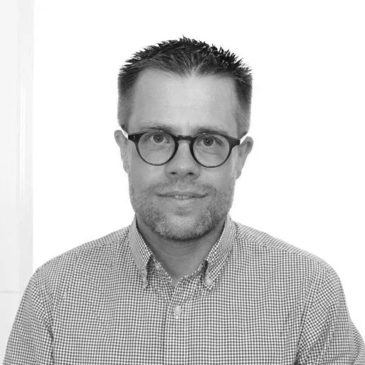 Black and white portrait of a man with short hair, wearing glasses, and a checkered button-up shirt, smiling slightly.