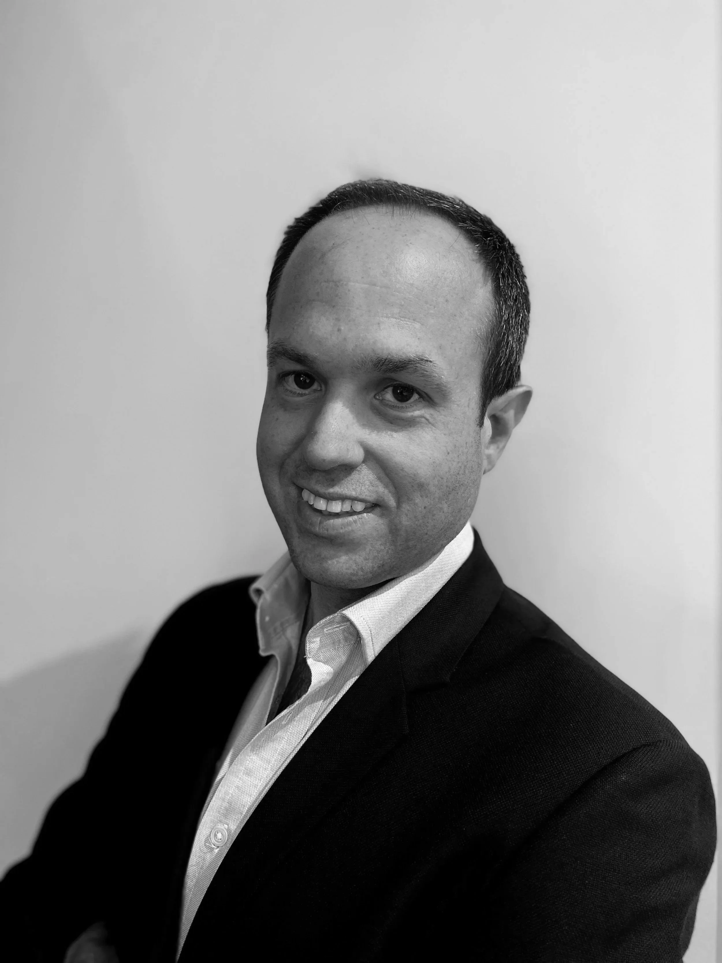 Black and white photo of a smiling man with short dark hair, wearing a dark blazer over a light-colored shirt, standing against a plain light-colored wall.