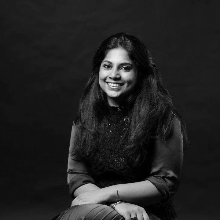 Black and white portrait of a smiling woman with long hair, wearing a dark, lace top and sitting against a dark background.