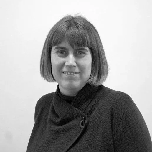 Black and white portrait of a woman with shoulder-length hair and bangs, smiling, wearing a dark top with a high collar, standing against a plain background.