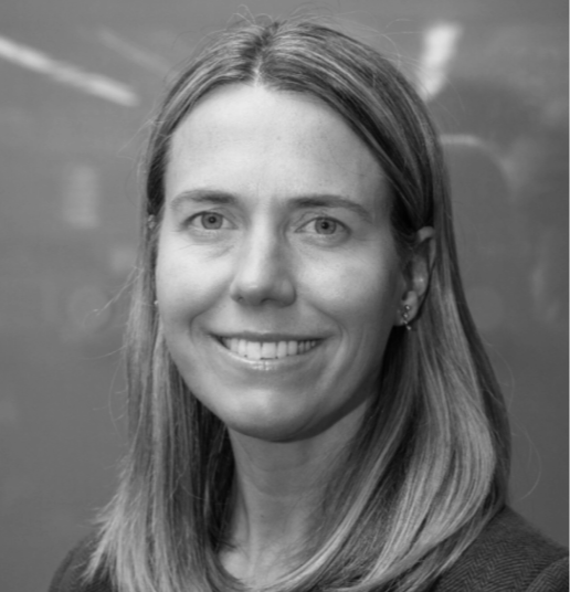 Black and white portrait of a woman with shoulder-length hair smiling, standing in front of a chalkboard.