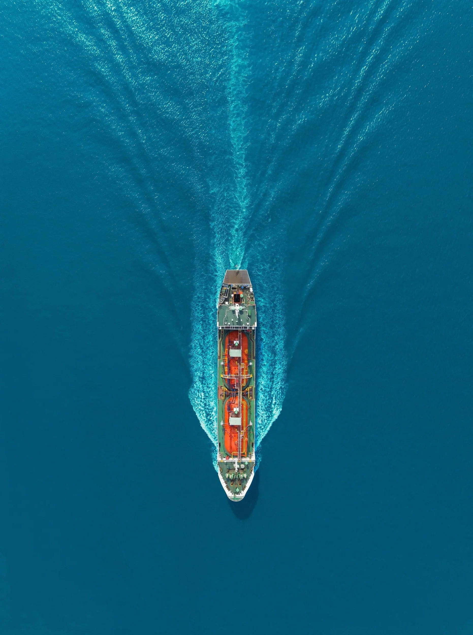 An aerial view of a large ship sailing through clear blue water, creating a wake behind it.