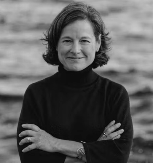 Black and white photo of a woman with short, wavy hair smiling, wearing a turtleneck sweater, standing outdoors with blurred water or shoreline in the background.