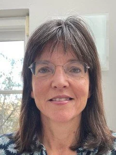 A woman with shoulder-length brown hair, wearing glasses and a blue patterned top, smiling in an indoor setting with a window in the background.
