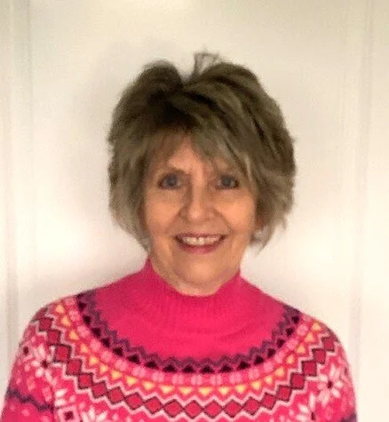 A smiling woman with short, layered hair wearing a colorful pink, red, black, yellow, and white patterned sweater.
