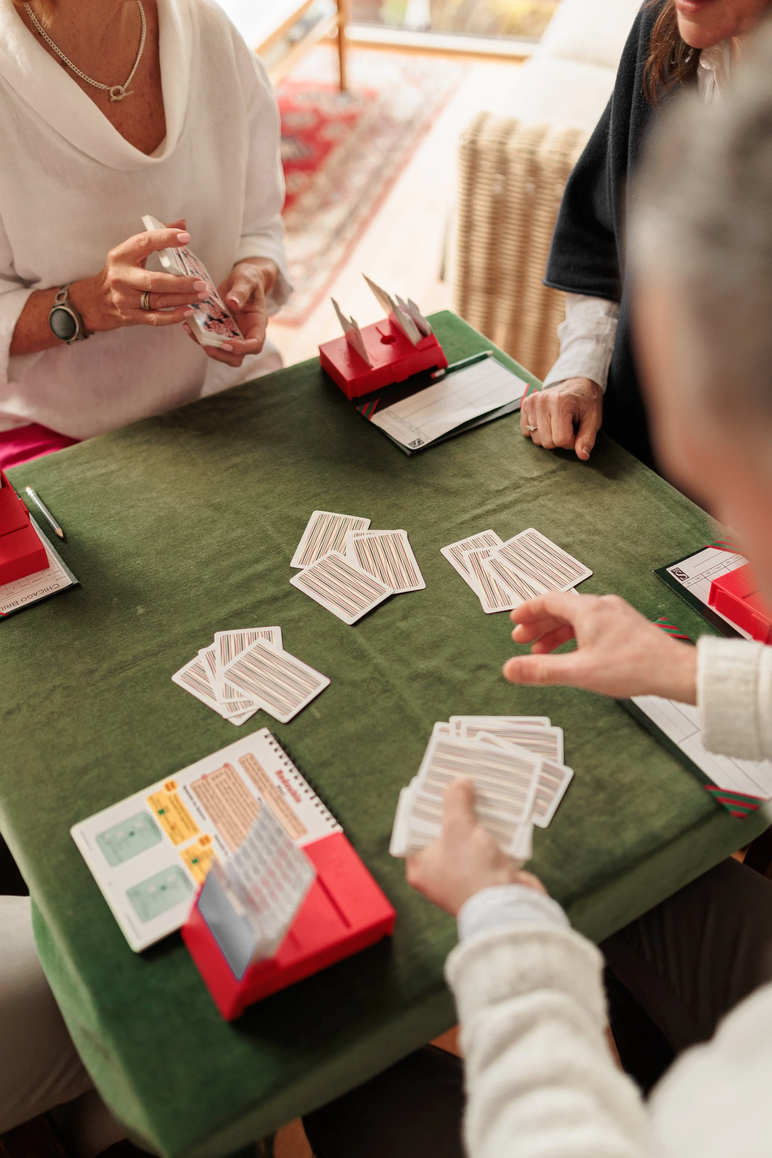 Four people playing a game of Rummikub around a green table indoors, with cards and score pads visible.