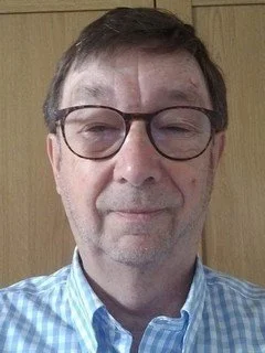 A man wearing glasses and a blue checkered shirt, standing in front of a wooden background.