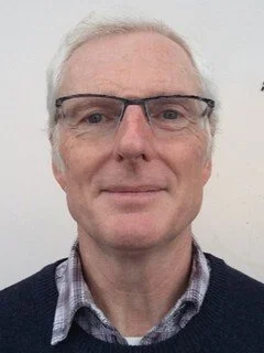 A middle-aged man with glasses, short gray hair, and wearing a collared shirt with a sweater, standing against a plain light background.