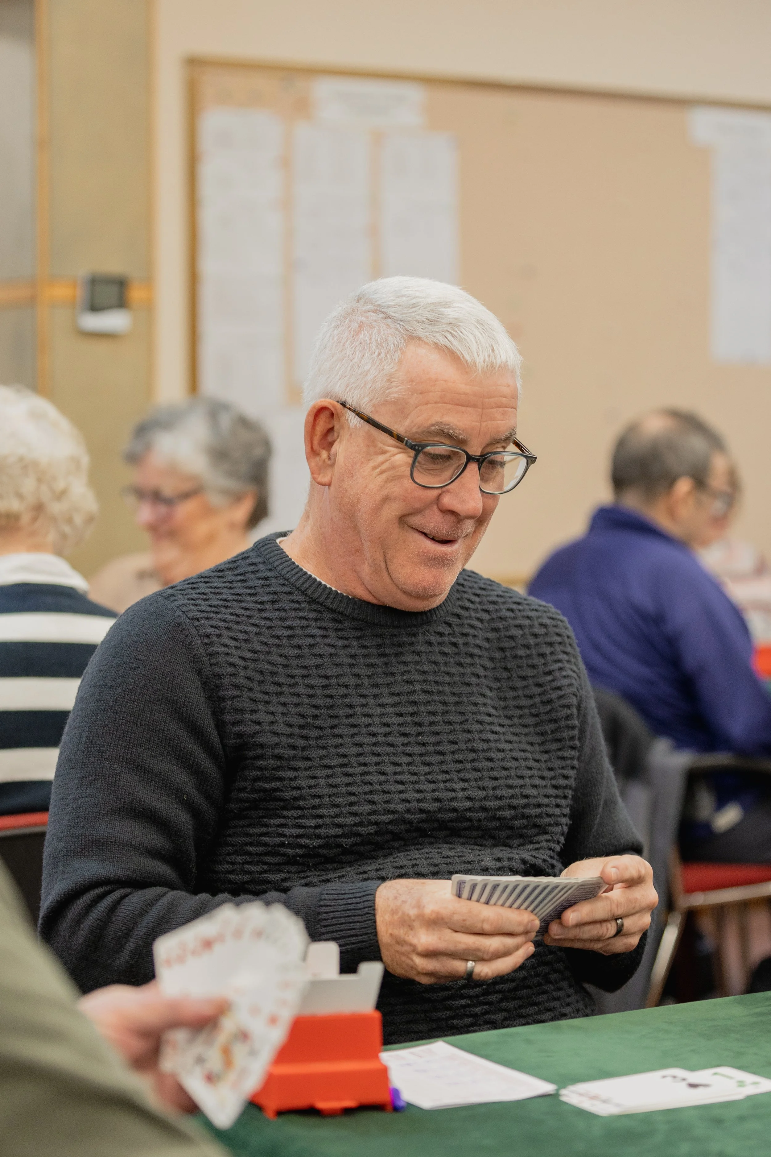 An older man with white hair and glasses smiling while playing a card game at a social gathering.