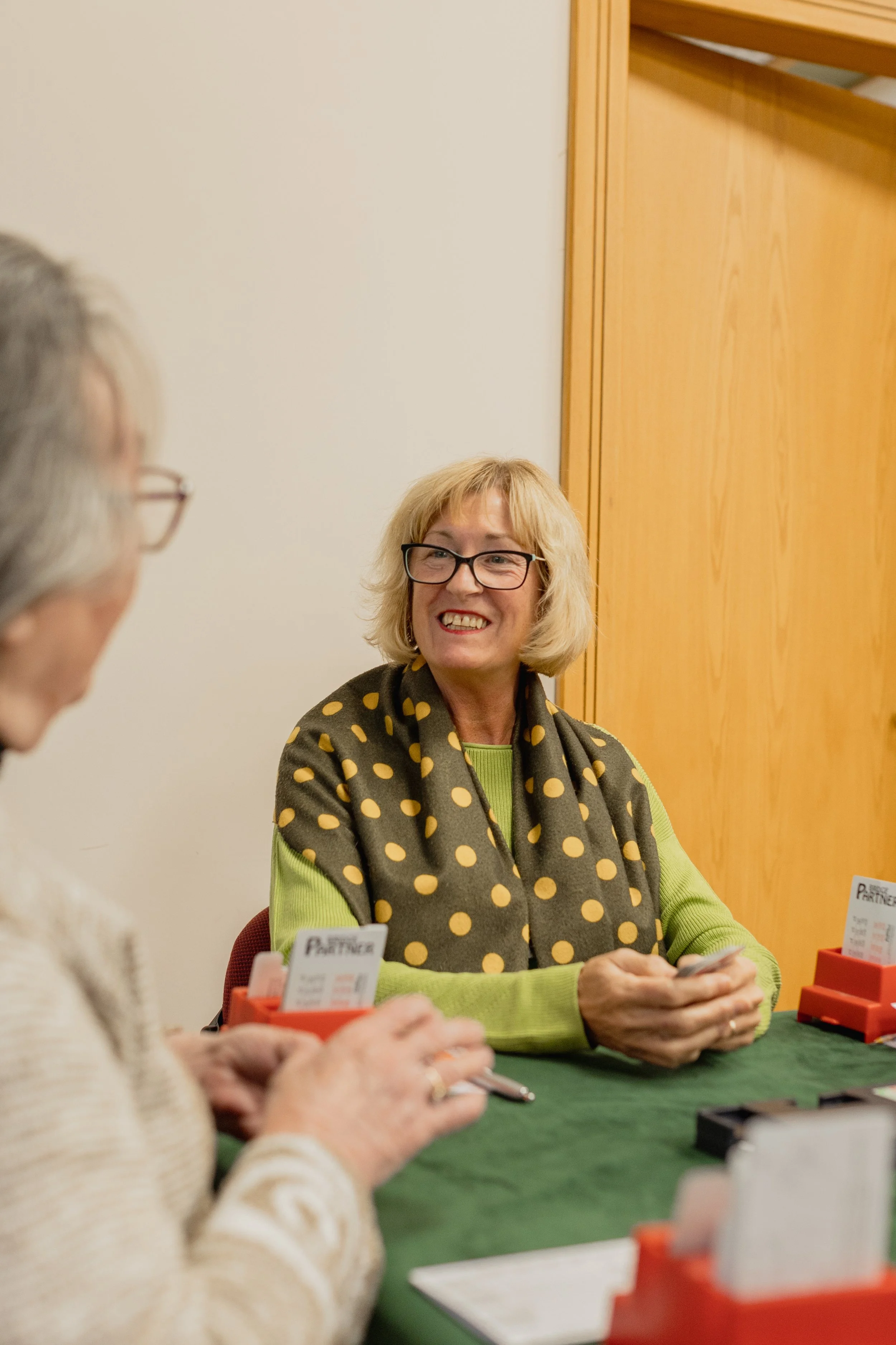 A woman with blonde hair and glasses, smiling, sitting at a table with another woman, in a room with wooden walls.