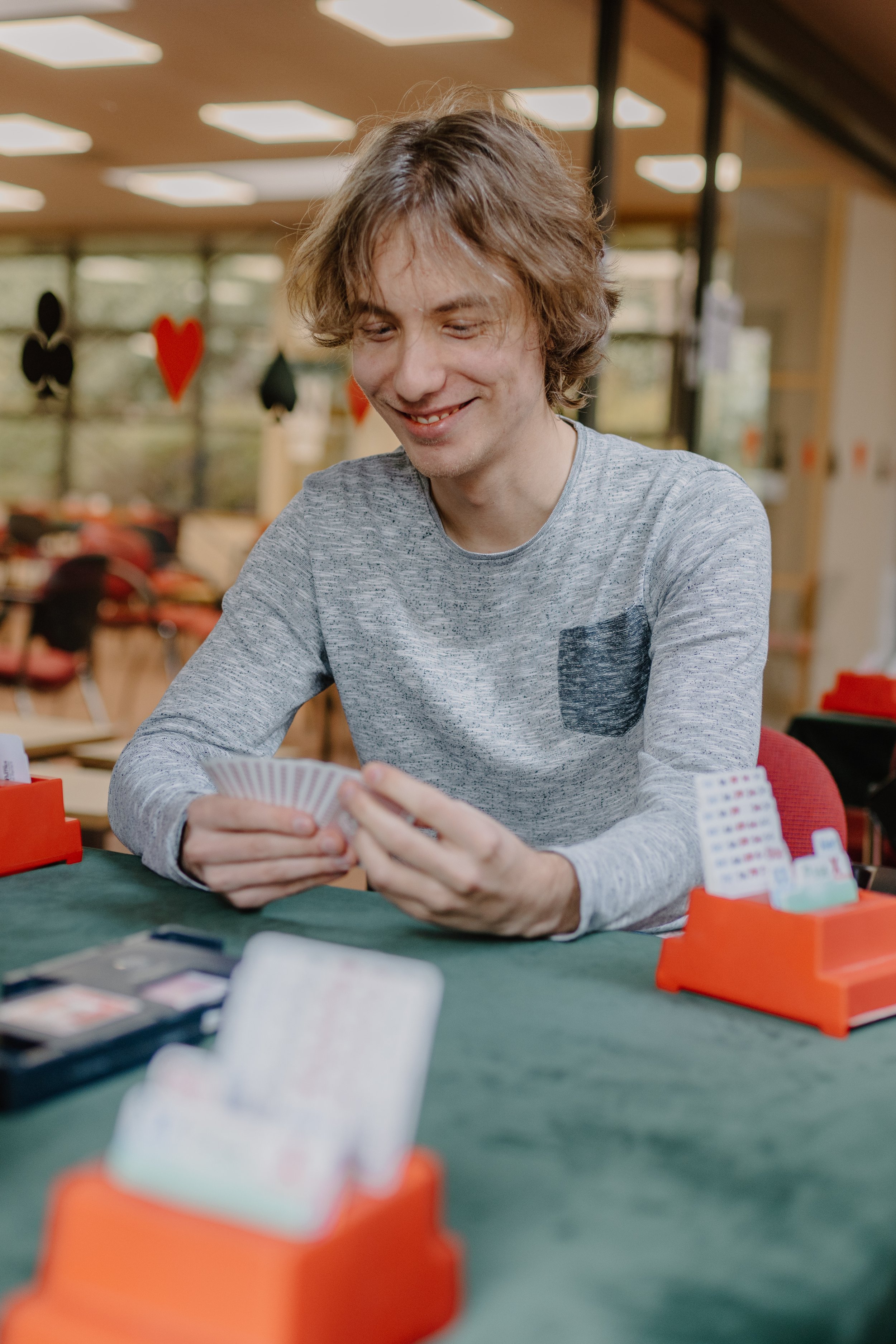 Young man with wavy light brown hair and fair skin smiling while playing a card game at a table in a well-lit indoor setting, with playing cards and poker chips visible on the table