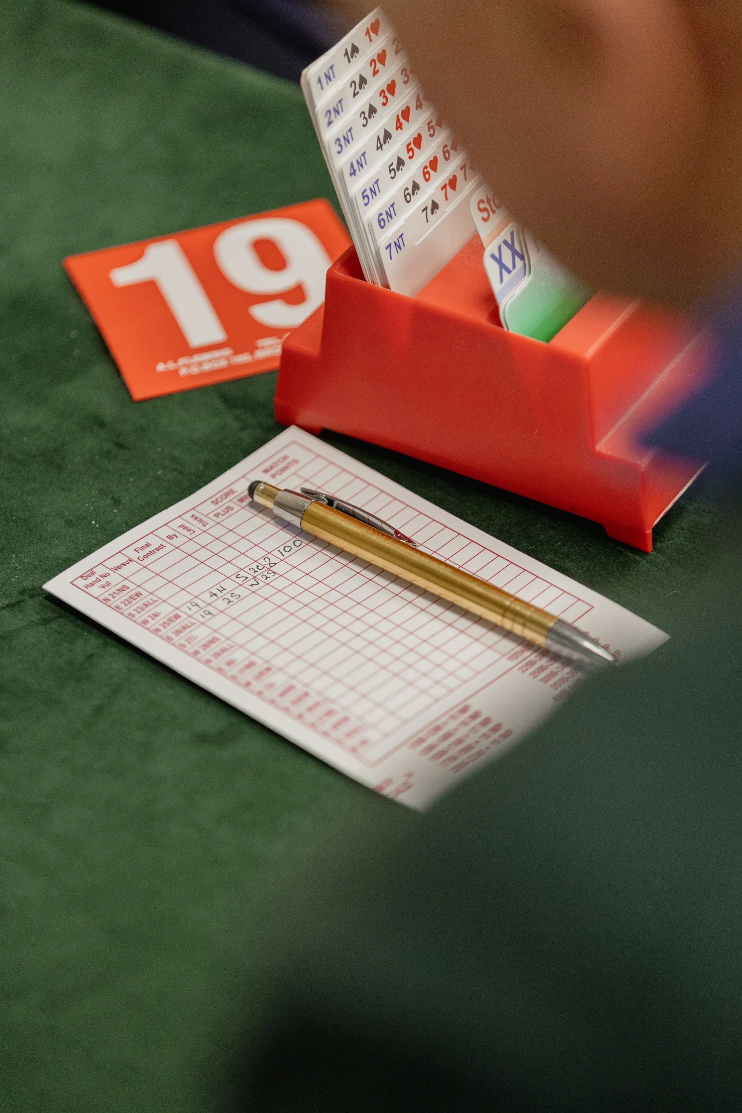 A betting slip with a gold pen on it, a red and white bingo card, a red number 19 sign, and a red ticket rack with poker cheat sheets in it, all on a green felt surface.