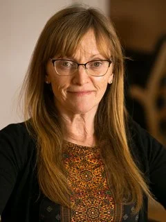 A woman with long, straight, light brown hair and glasses, wearing a dark top with a patterned shirt underneath, smiling at the camera.