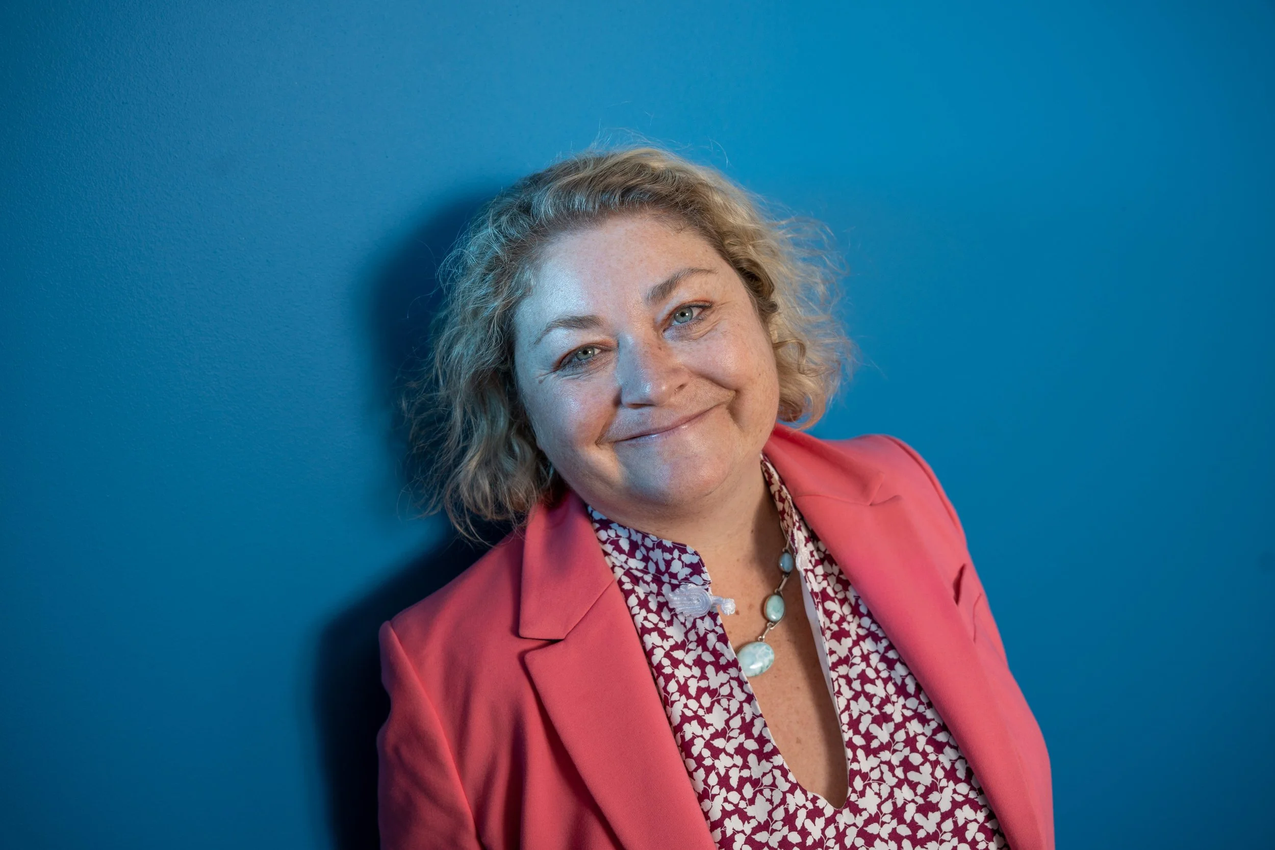 A woman with curly blonde hair and blue eyes smiling, wearing a pink blazer over a patterned blouse, standing against a solid blue wall.