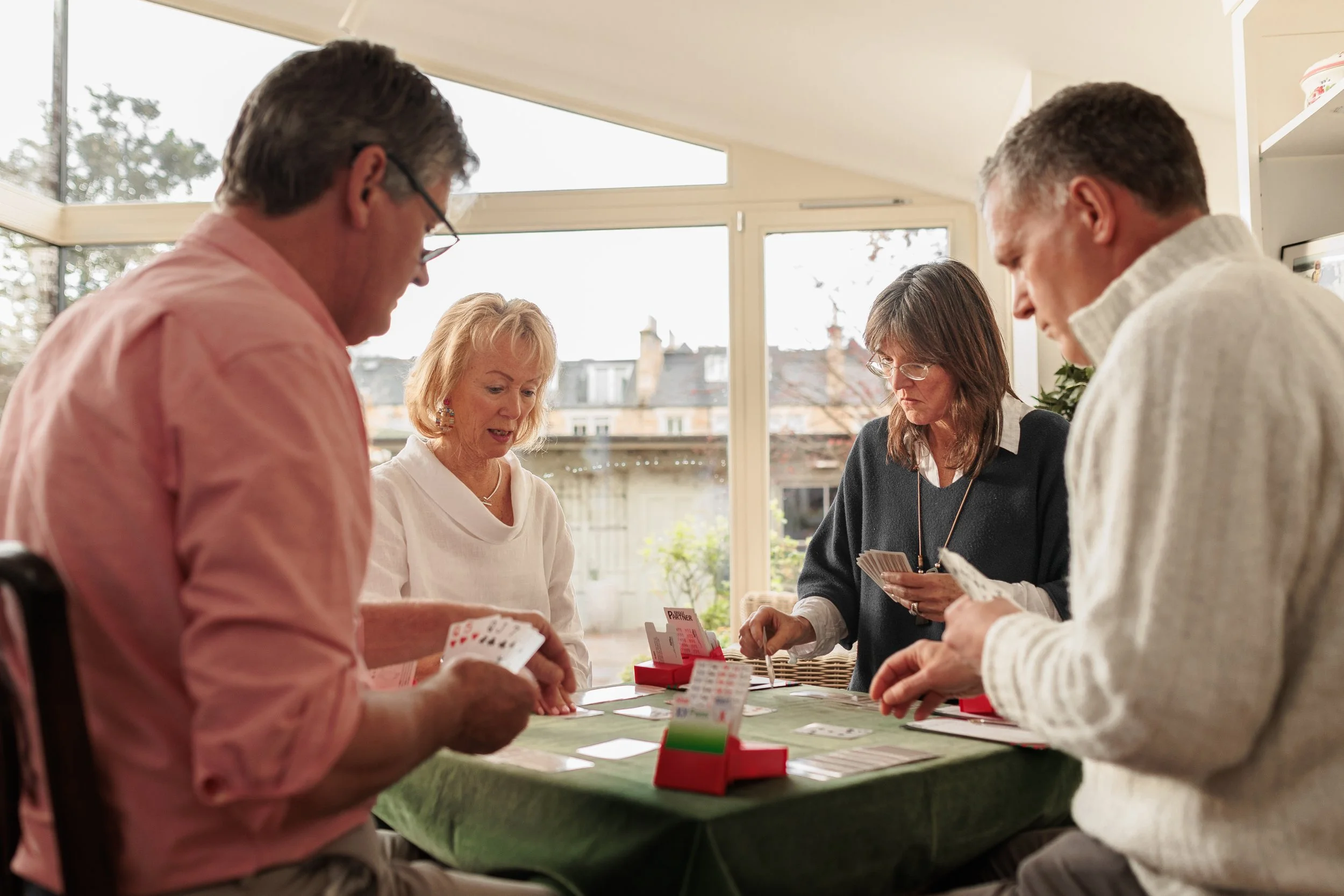 Four adults playing a card game around a green table in a well-lit room with large windows.