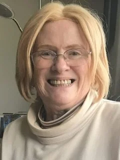 A smiling woman with blonde hair, wearing glasses and a white turtleneck, in an indoor setting.