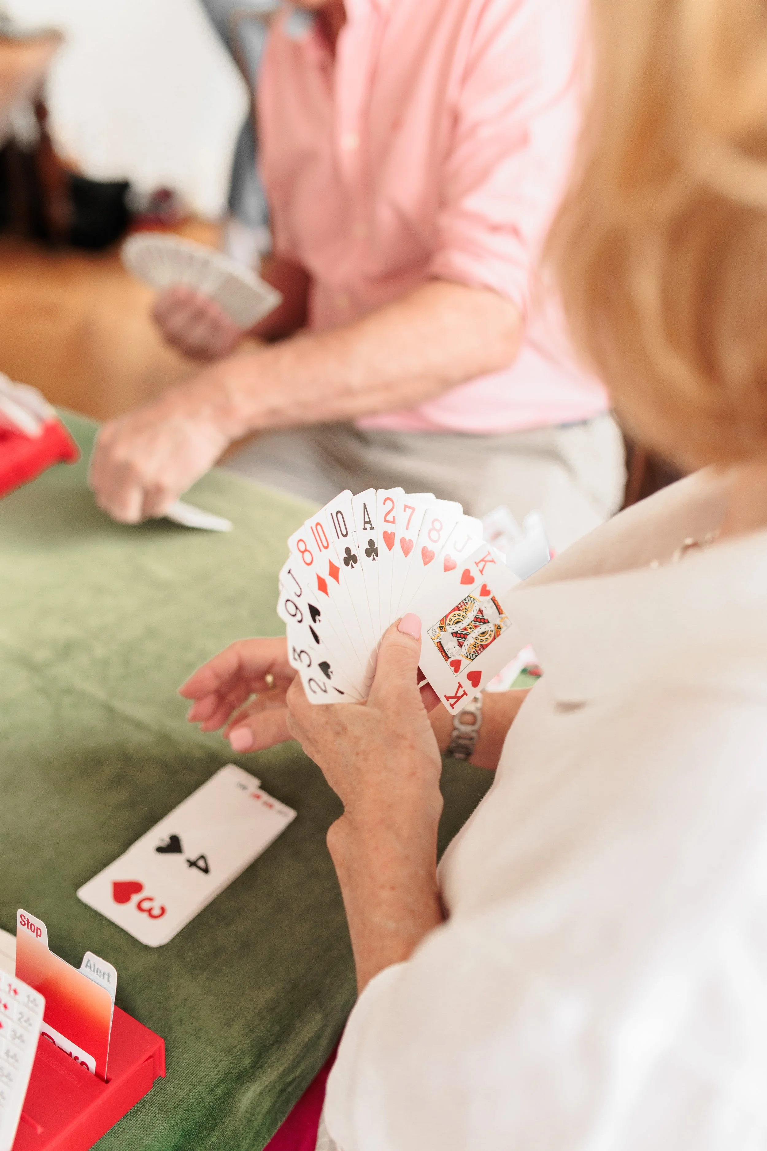 Two elderly women playing a card game at a green table, with playing cards visible in their hands and on the table, including a discarded card with a heart suit.