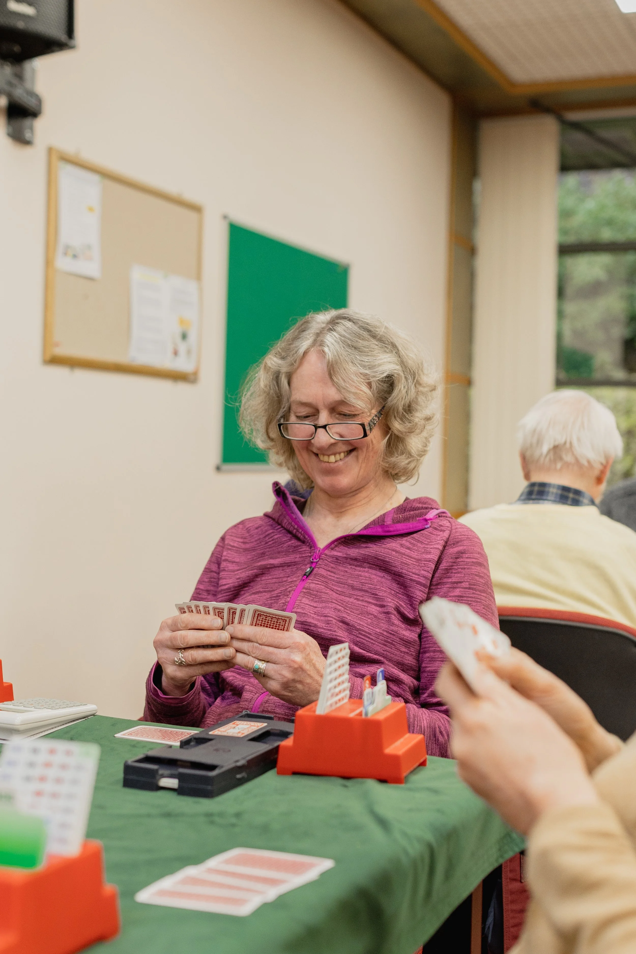 A smiling woman with curly blonde hair and glasses playing a card game at a green table in a room with bulletin boards on the wall. She is dressed in a purple hoodie and is holding playing cards. In the background, other people are also sitting and playing.