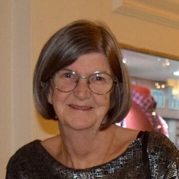 A woman with short brown hair, wearing glasses and a black sequin top, smiling indoors.