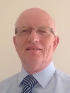 A man with glasses, wearing a light blue shirt and a striped tie, smiling slightly against a plain background.