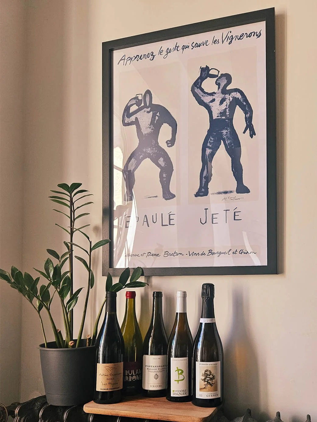 Une affiche artistique montrant deux hommes nus, l'un en train de jeter un objet et l'autre en position de lancer, avec du texte en français. En dessous, plusieurs bouteilles de vin sont placées devant un pot de plante.