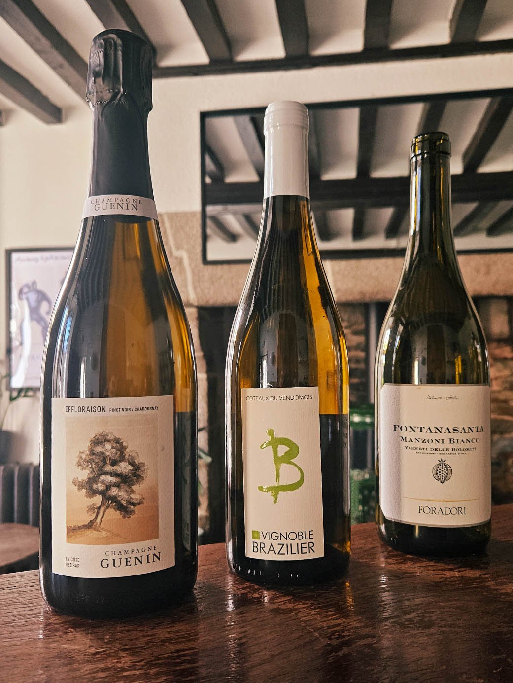 Trois bouteilles de vin alignées sur une table en bois dans une pièce avec plafond en poutres apparentes et cheminée en arrière-plan.