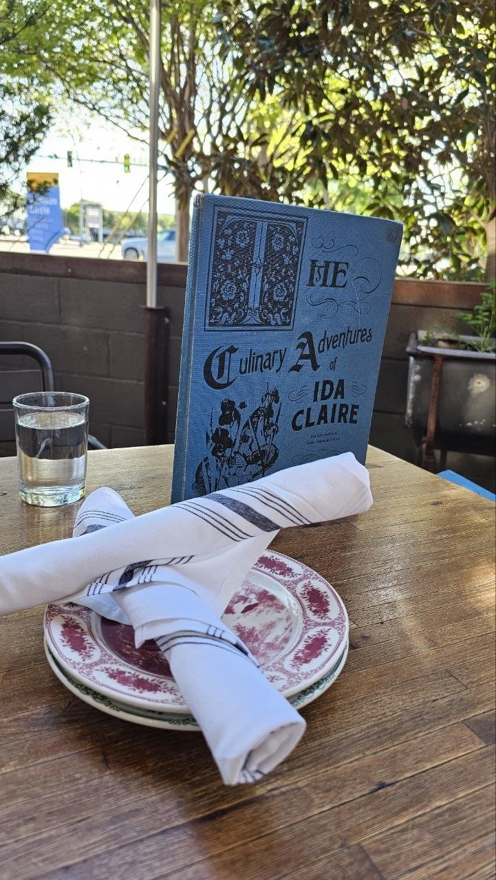 A table setting with a glass of water, a plate with a napkin, and a blue menu book titled "The Culinary Adventures of Ida Claire" inside a restaurant with outdoor seating and greenery.