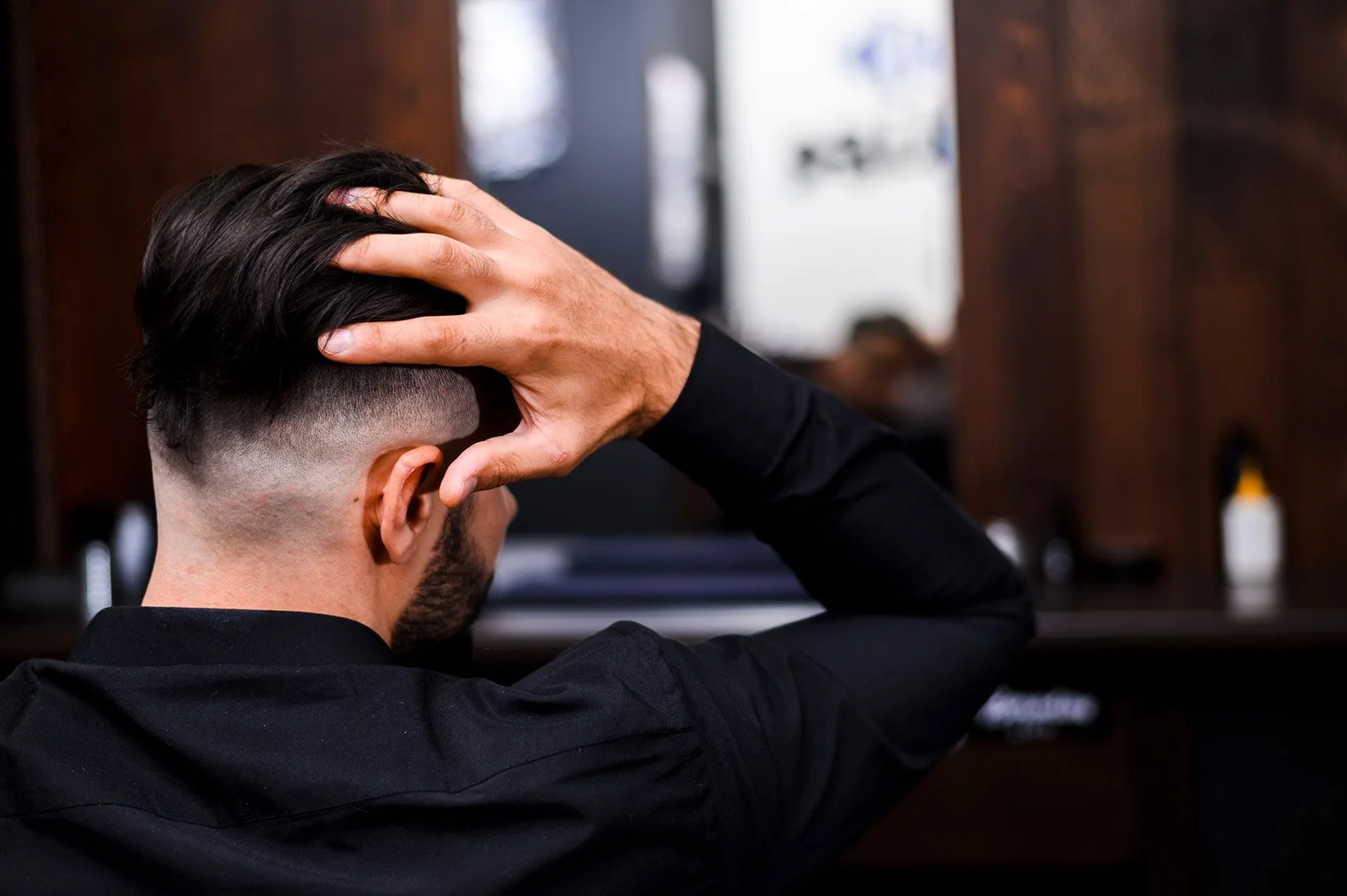 Man with a fade haircut sitting in a barber shop, touching his head, with a mirror and barber supplies in the background.