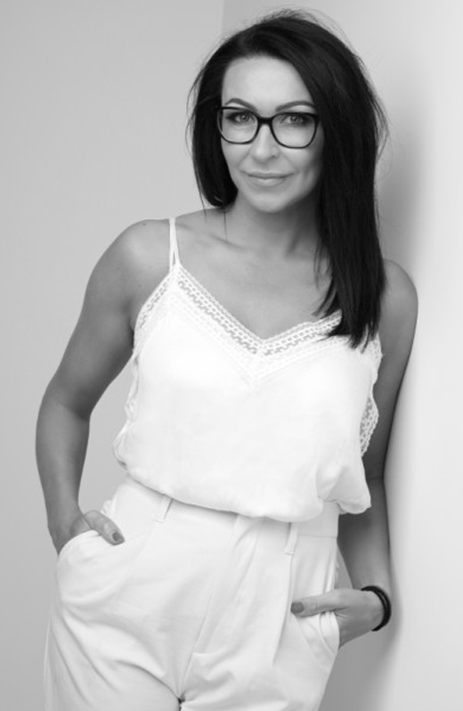 A woman with dark hair and glasses wearing a white sleeveless blouse and white pants, standing and posing against a light-colored background.