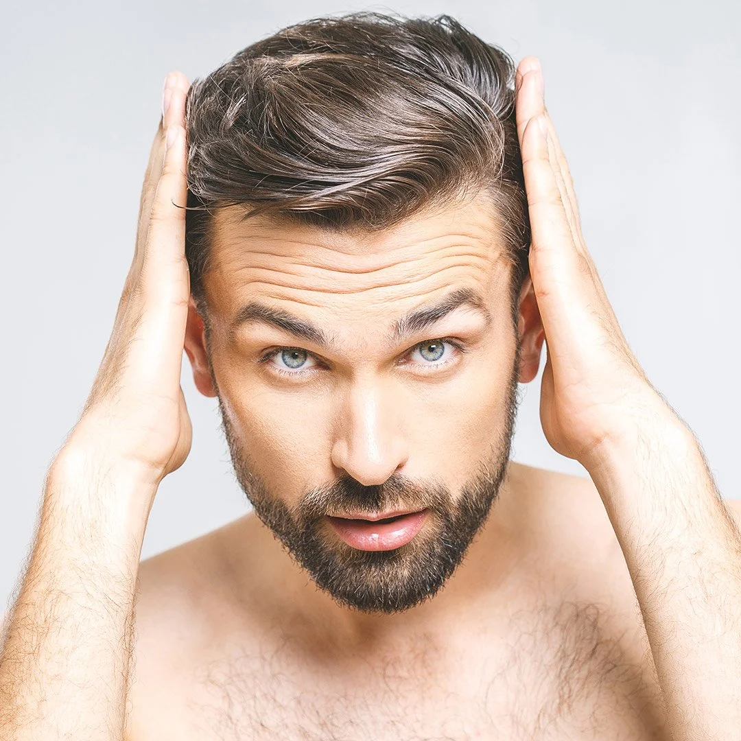A shirtless man with a beard and blue eyes holding his hands to his head, looking surprised or startled.