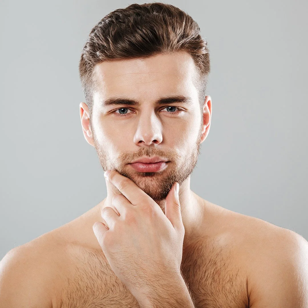 A shirtless young man with light skin, brown hair, and a beard, touching his chin thoughtfully, against a plain gray background.