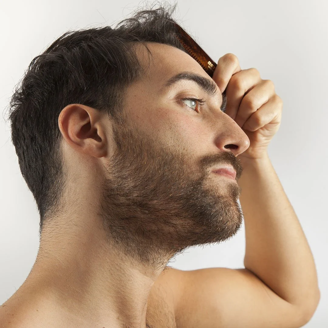 A shirtless man with dark hair and a beard holds a brown comb to his head, looking upwards with a neutral expression.