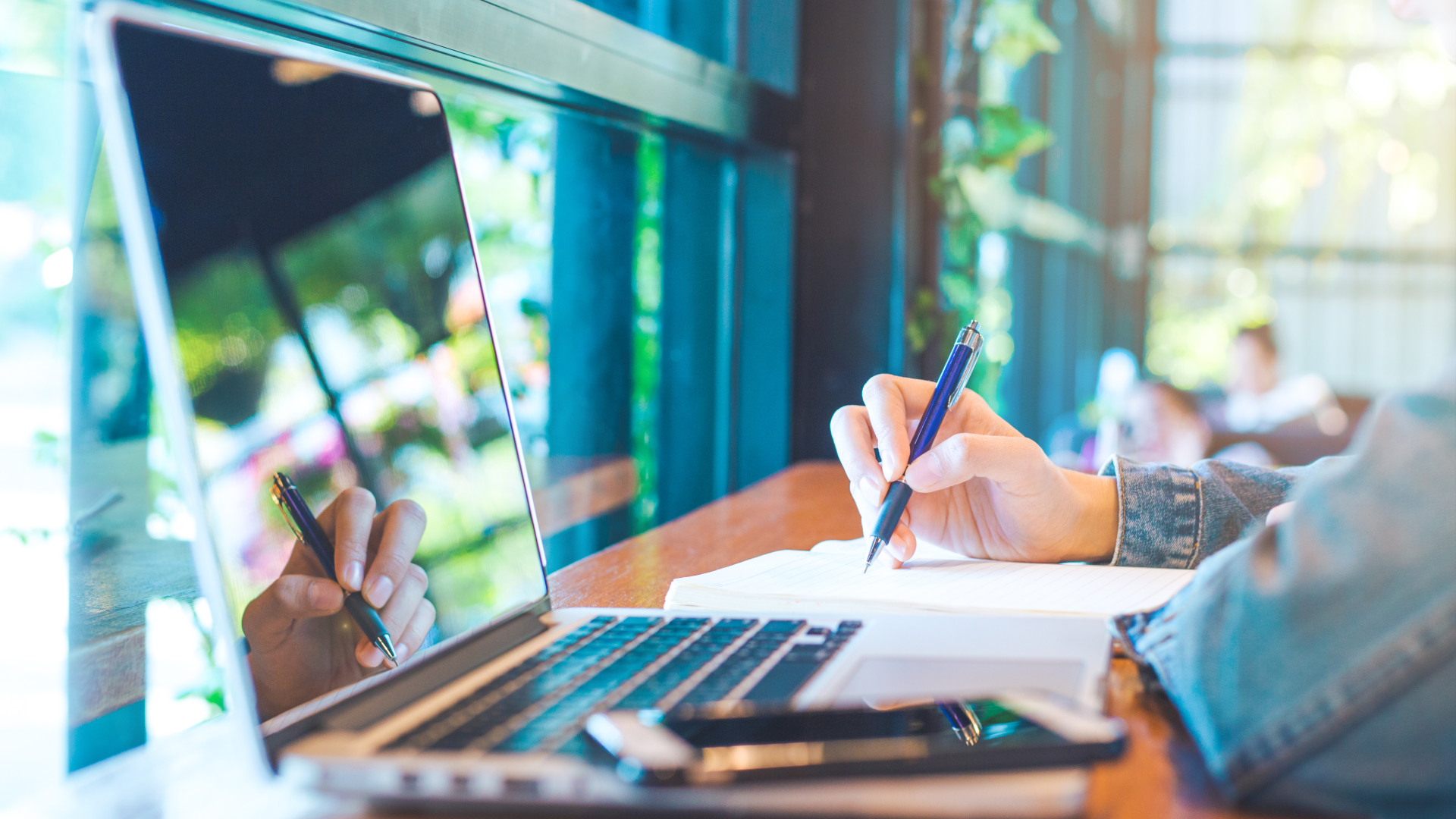 A person sitting at a wooden table working with a laptop, notebook, and pen, outdoors in a bright, green environment with sunlight filtering through trees.