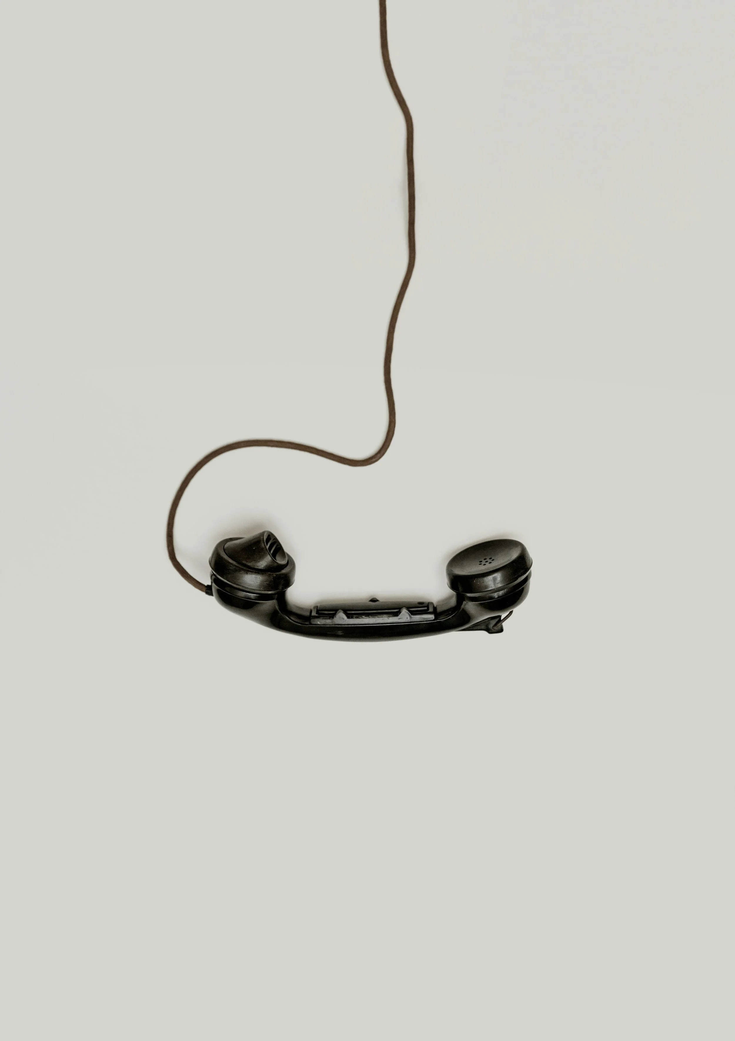 Old black telephone handset connected to a wire hanging against a plain light background.