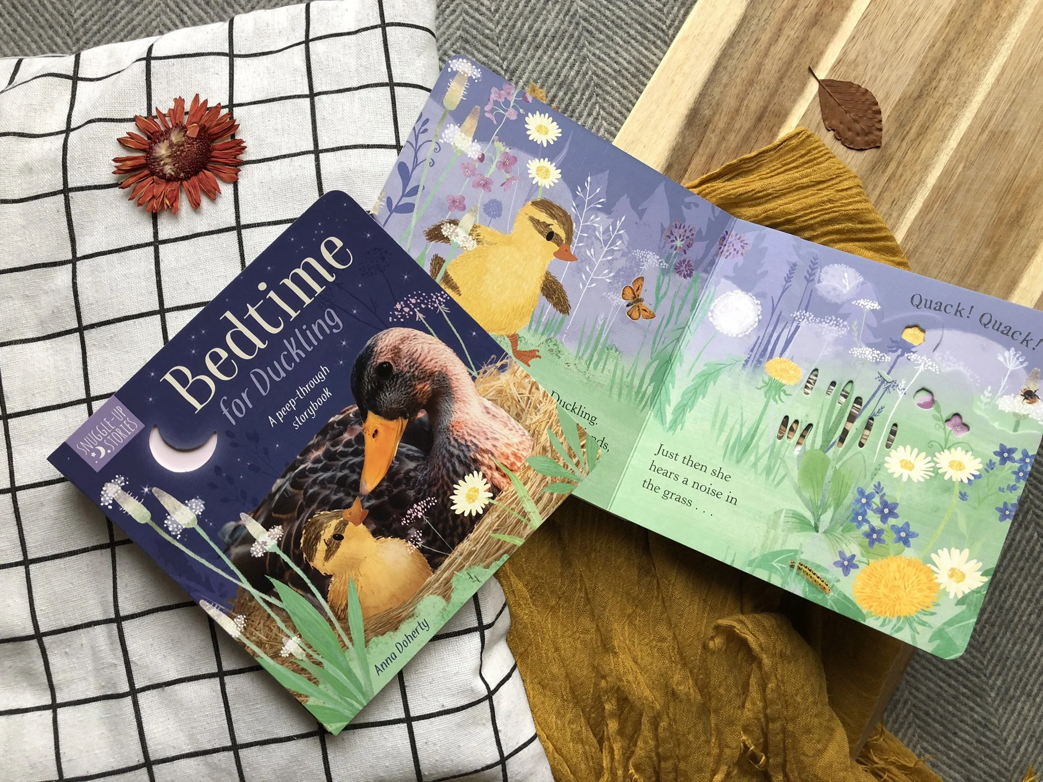 Children's book titled 'Bedtime Duckling' resting on a checkered cloth with a dried leaf and mustard-colored cloth beside it. The book features an illustration of a duckling in a natural setting.