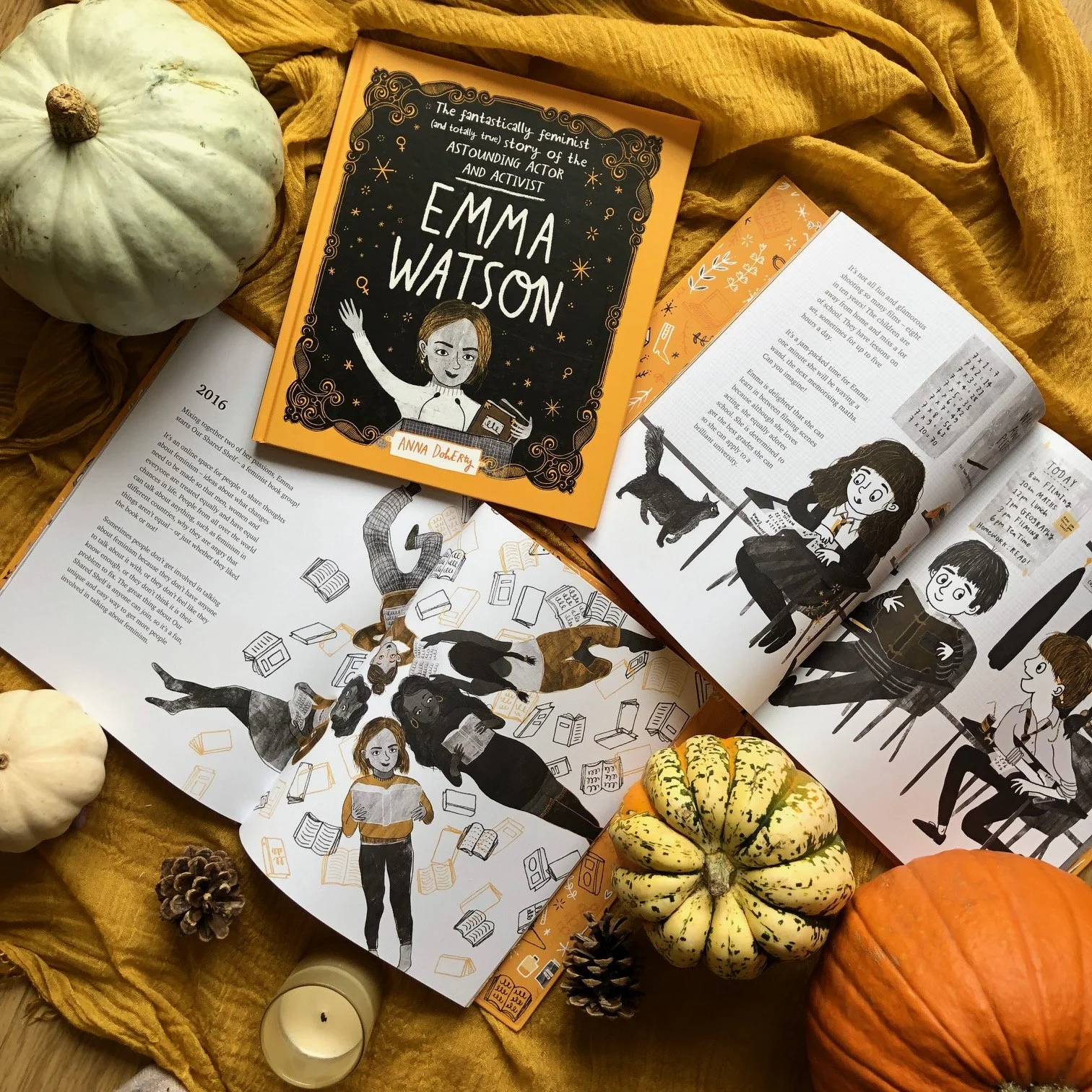 Open books and illustrations surrounded by pumpkins, a pine cone, a candle, and a yellow cloth.
