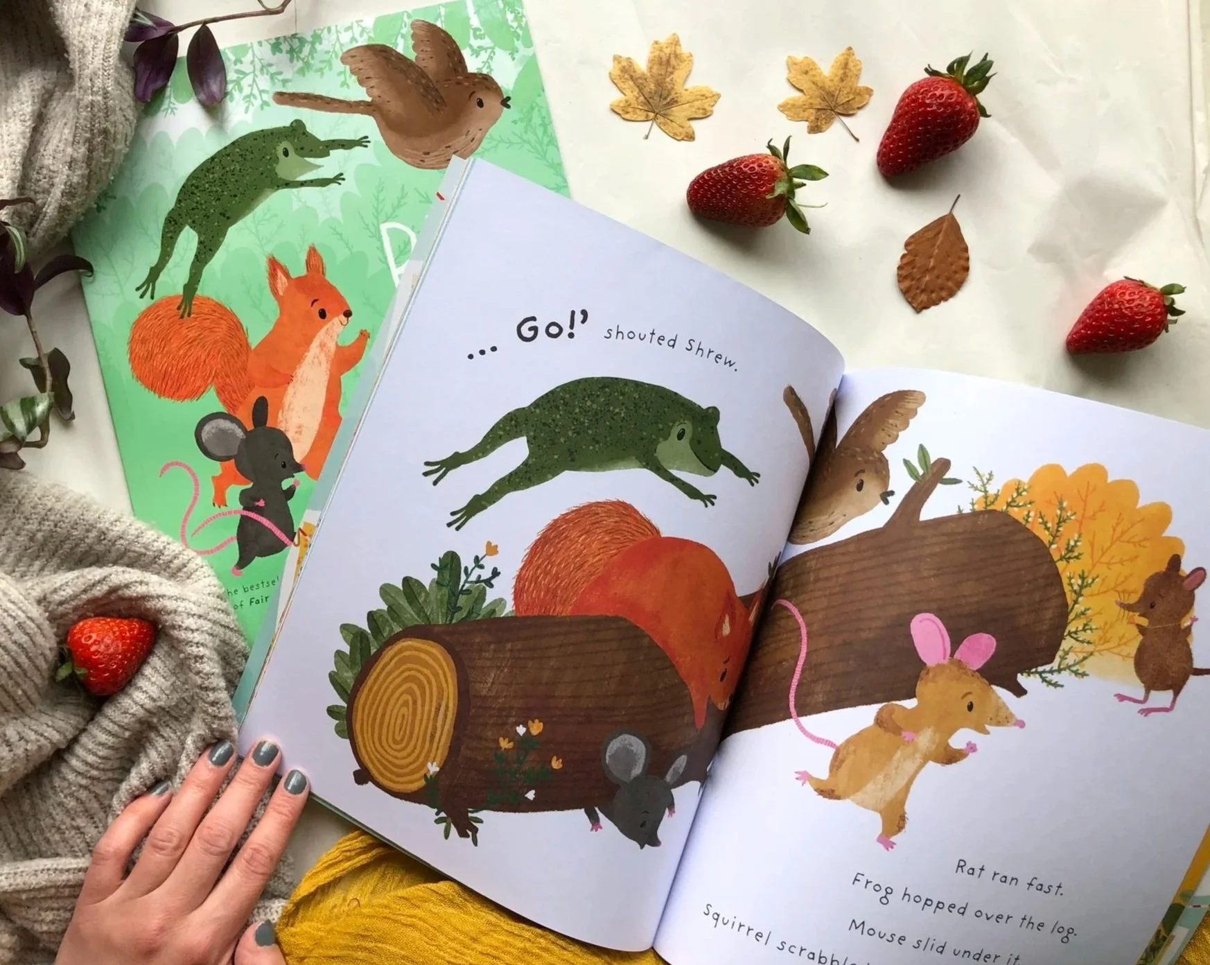 Open children's book with illustrations of woodland animals and forest scene, surrounded by strawberries, leaves, and a knitted fabric with a strawberry decoration.