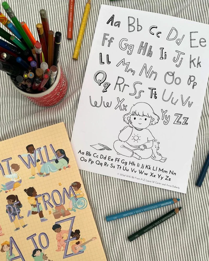 A children's alphabet learning sheet with uppercase and lowercase letters, a girl drawing, colored pens in a cup, colored pencils, and a notebook with drawings of children and the alphabet, all on a striped fabric surface.