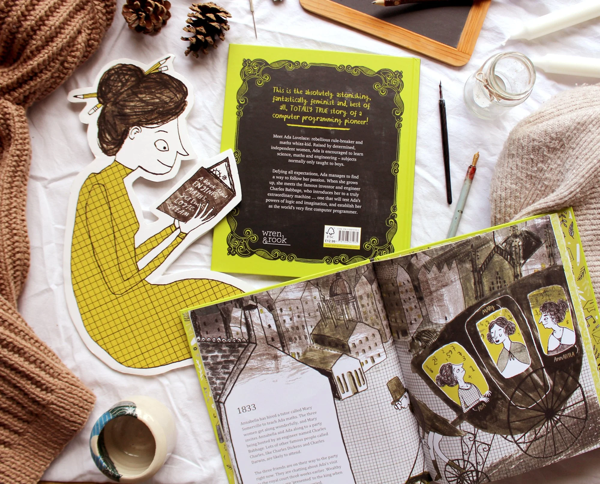 A flat lay of various items including a yellow book with black writing, a sticker of a woman reading, pinecones, a small chalkboard, a mason jar, a small paintbrush, a knitted blanket, and other miscellaneous items on a white cloth.