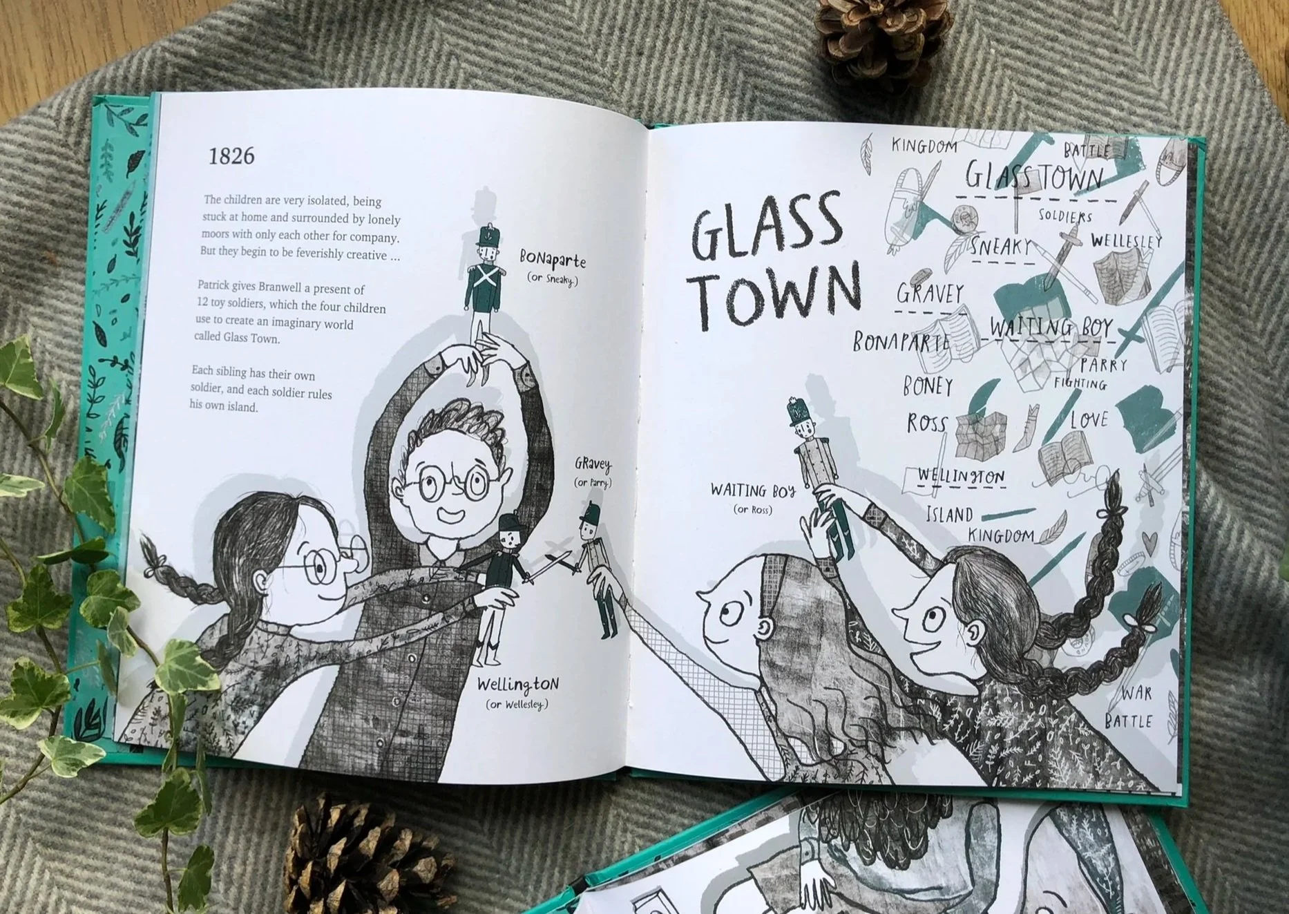 Open coloring book titled 'Glass Town' featuring black and white illustrations of children dressed as soldiers, holding hands, and surrounded by words like 'Wellington,' 'Wellesley,' 'Boney,' 'Gravey,' 'Waiting Boy,' 'Parry,' and 'Love.' The pages are resting on a surface with pinecones, a sprig of ivy, and a striped fabric background.