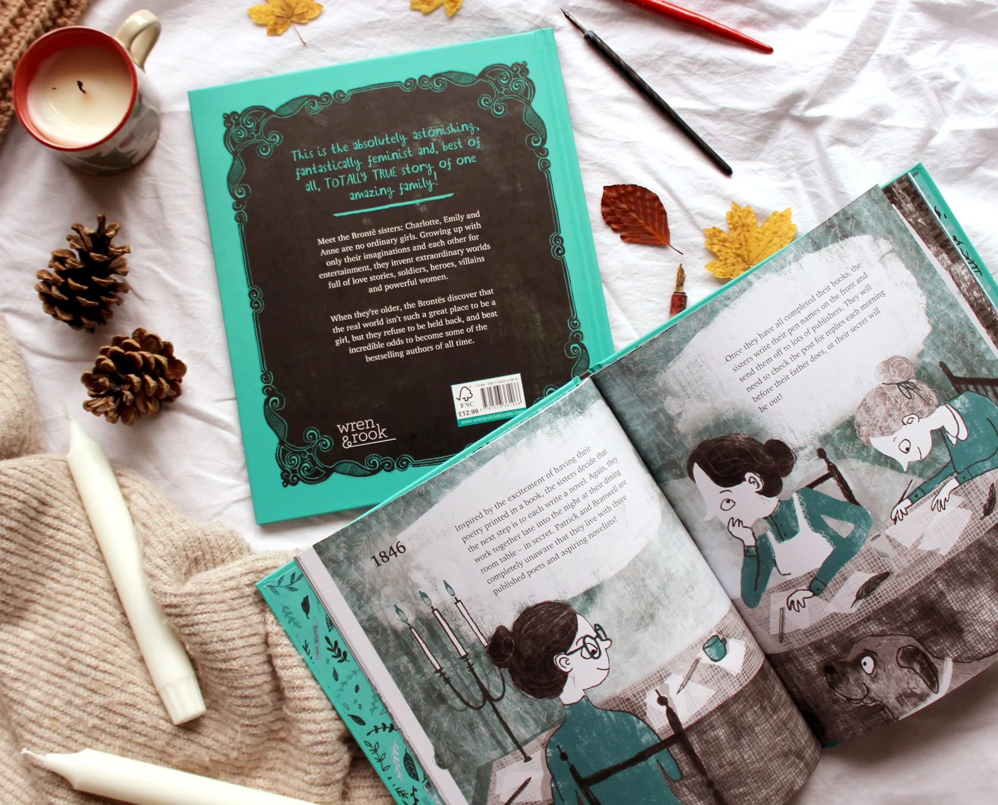 Open children's book with illustrations of children, surrounded by pine cones, autumn leaves, a candle, and fall-themed decorations on a white surface.