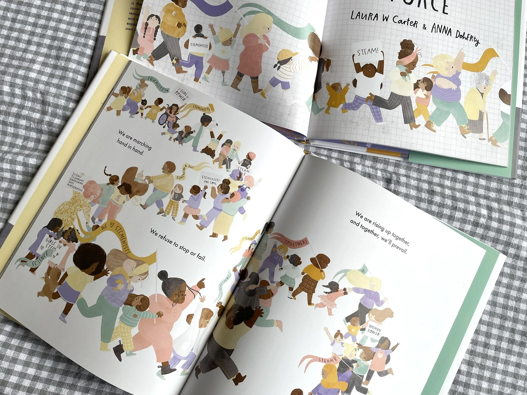 Open book pages showing illustrations of diverse children and adults marching and raising their hands with messages promoting equality, feminism, science, technology, engineering, arts, and mathematics, against a checkered fabric background.