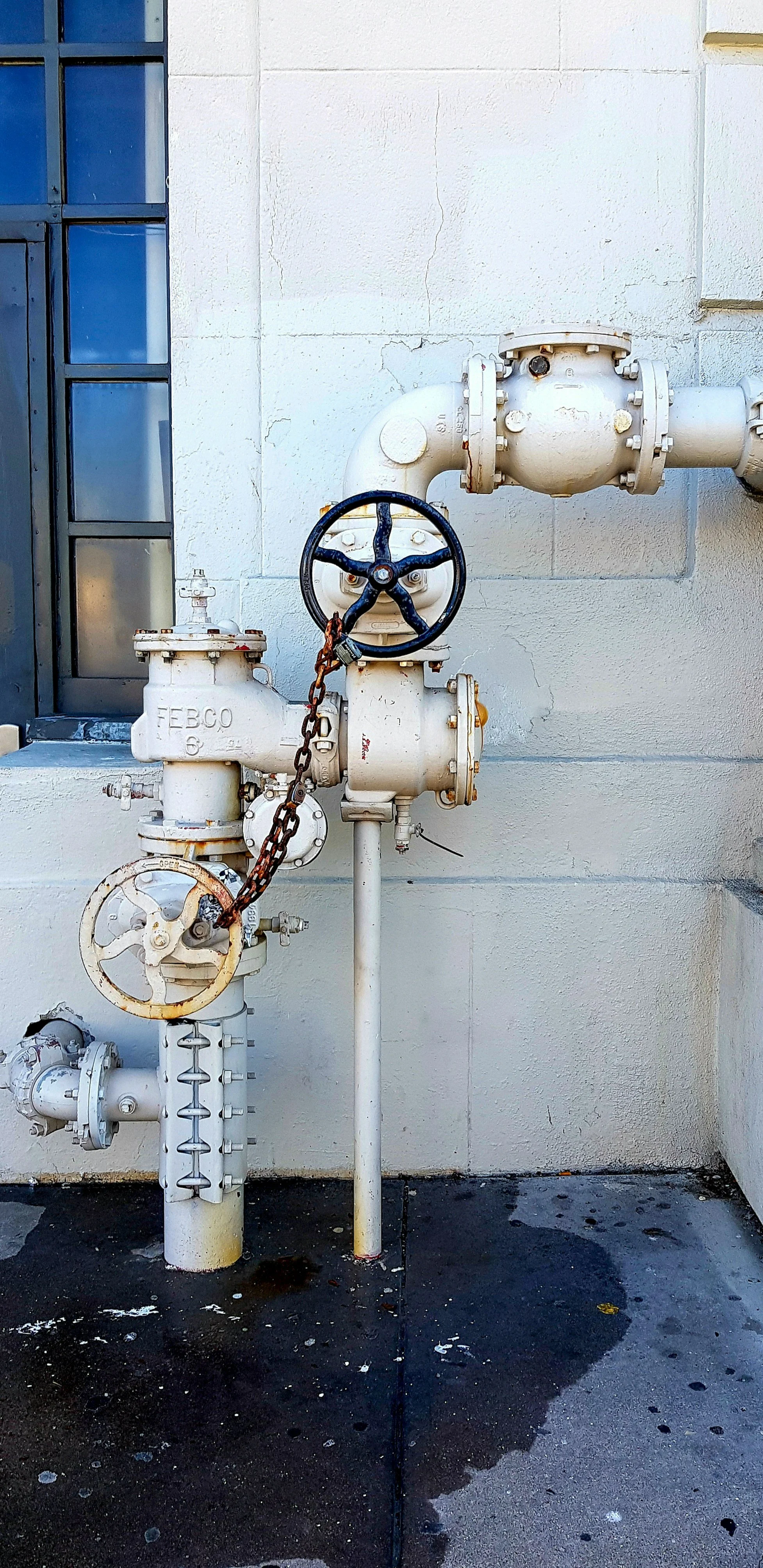 White industrial pipes and valves against a white wall, with a rusty chain and a black wheel handle on one valve.
