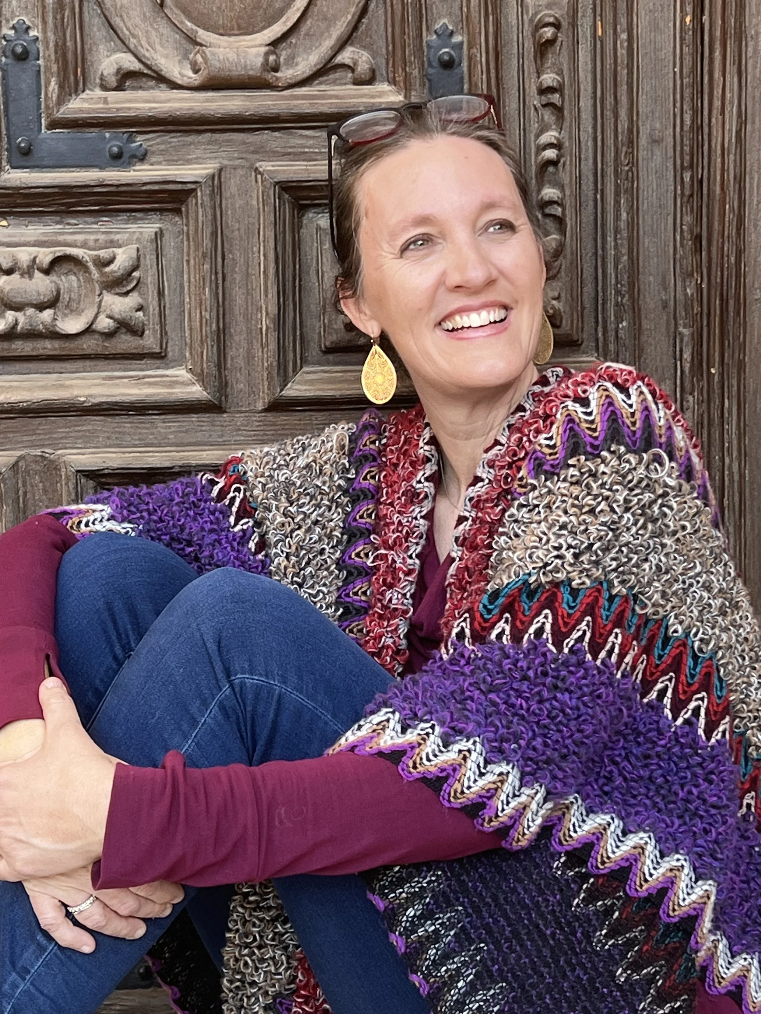 Close-up of a woman with short hair, smiling, wearing glasses on her head, gold earrings, and a multicolored, textured, patterned sweater. She is seated against a wooden door with carved details.