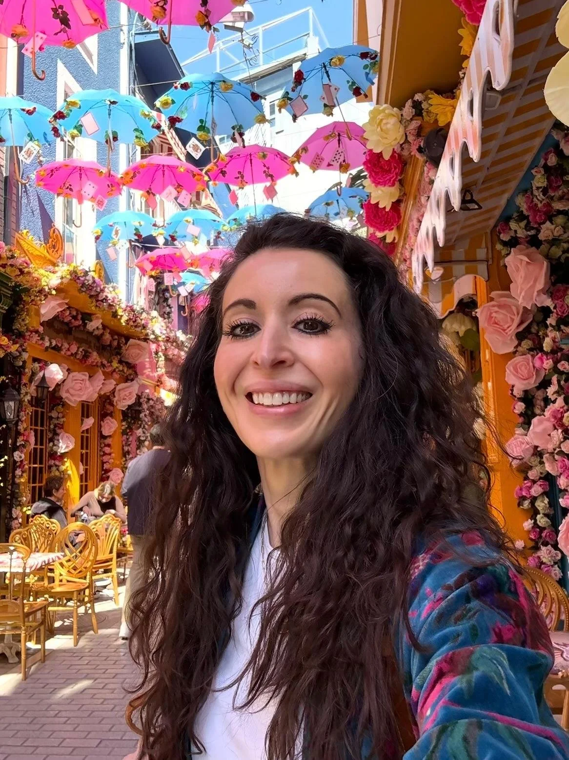 A woman with long curly dark hair taking a selfie in a vibrant outdoor setting decorated with pink and blue umbrellas hanging overhead, covered in flowers and surrounded by pink roses and yellow chairs.